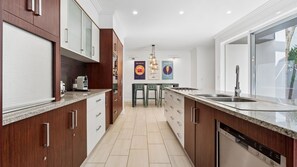 A modern kitchen with wooden cabinets, a granite countertop, a built-in dishwasher, and a central island with bar stools. - The North Point Mansion (Kingscliff)