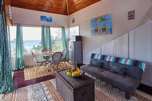 A room with a view of the ocean, a dining area with chairs and a table, a sofa with cushions, and a fruit basket.