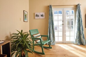 Une pièce avec un plancher en bois, un fauteuil à bascule doté d’un coussin à rayures, une plante en pot et une porte ornée de rideaux à motifs.