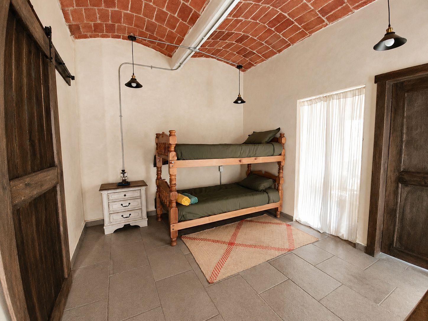 Una cama litera con estructura de madera, una mesita de noche pequeña, una lámpara y una ventana con cortinas.