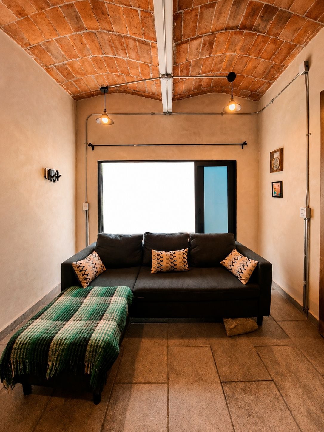 Una habitación acogedora con techo de ladrillo, un ventanal grande, un sofá negro con cojines estampados y una manta verde y blanca a rayas sobre el suelo.