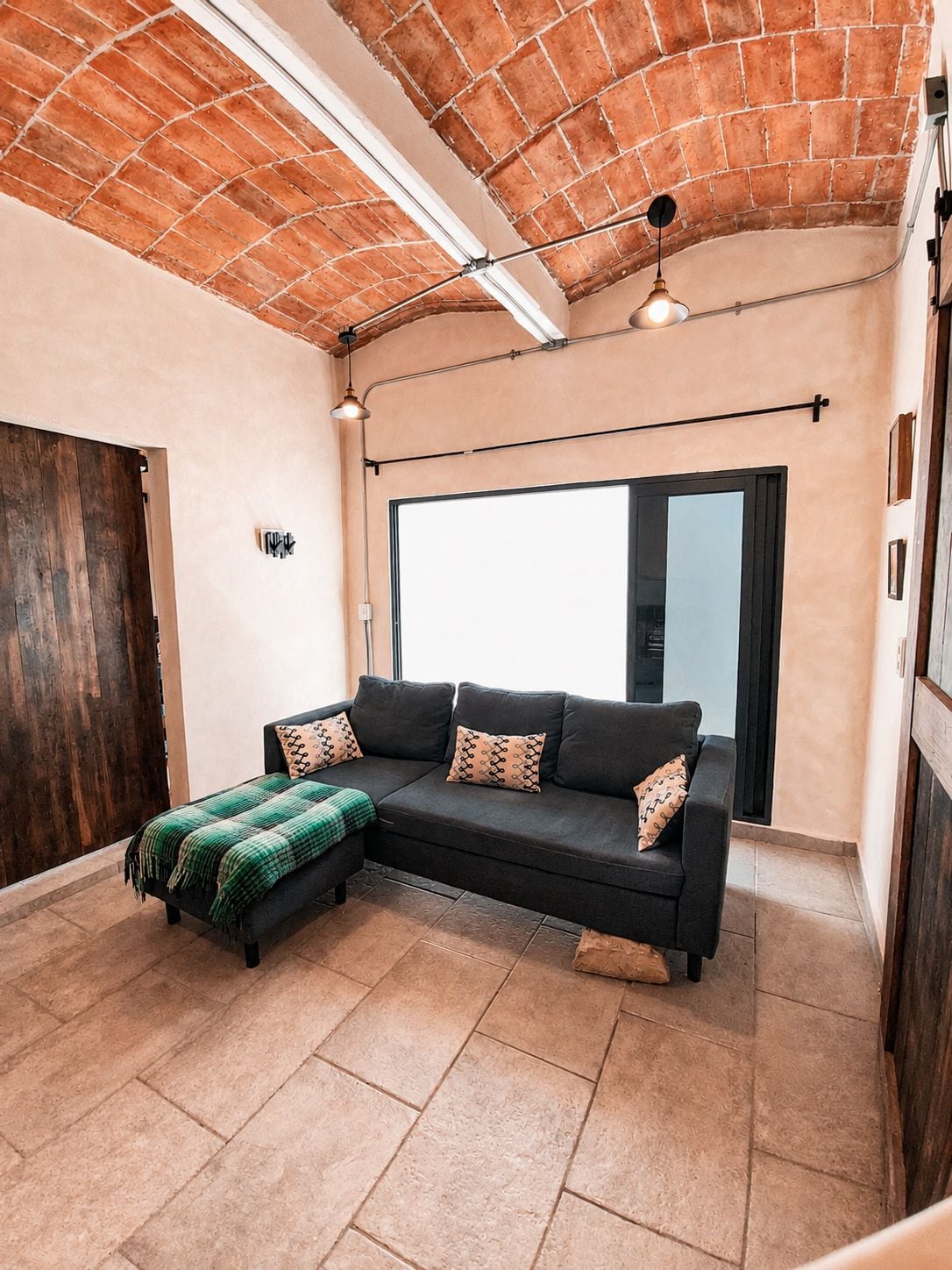 Una sala de estar moderna con un sofá modular negro, una manta verde y un ventanal que proporciona luz natural.