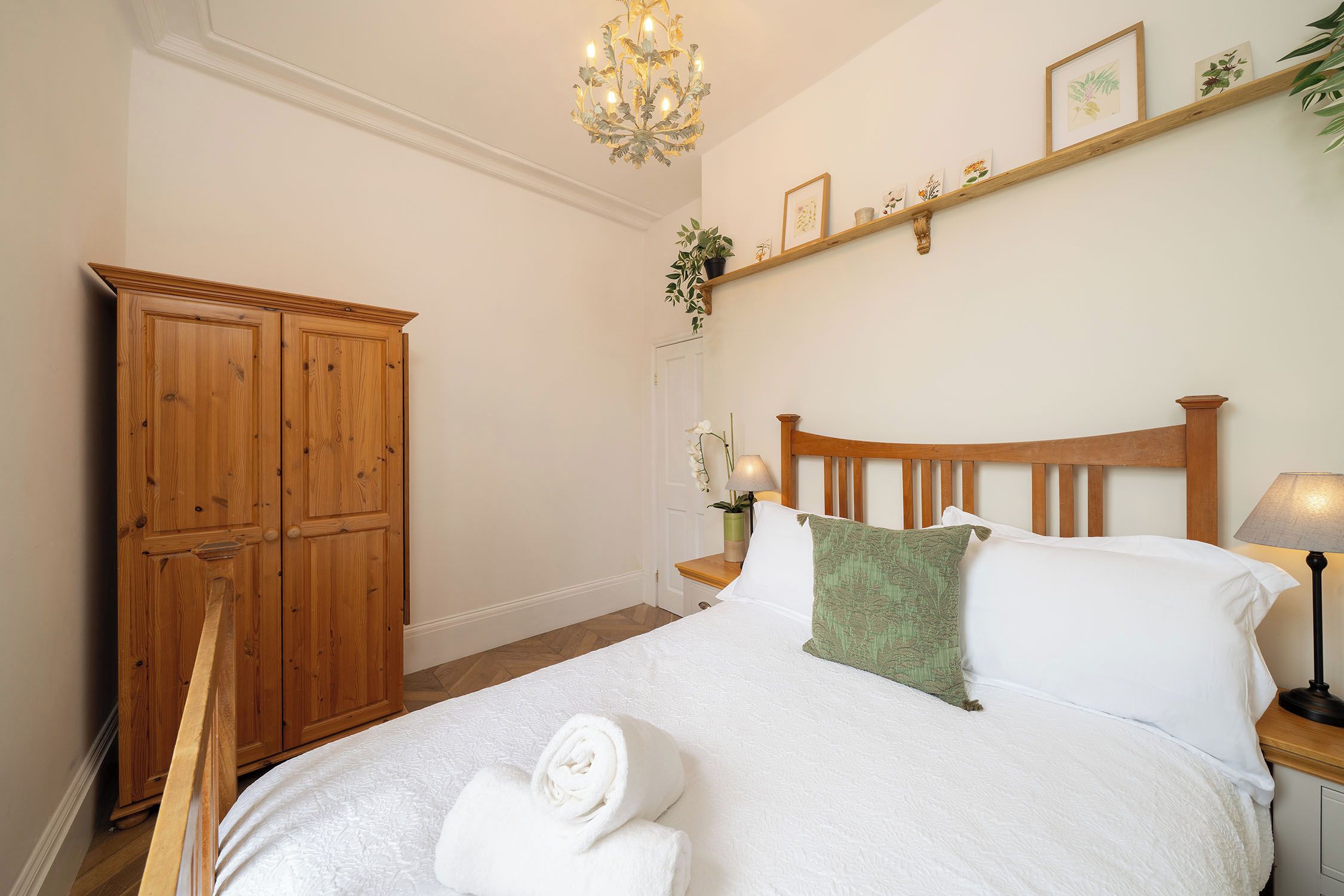 A bedroom with a wooden bed, white bedding, a chandelier, a wooden wardrobe, and a shelf with framed pictures.