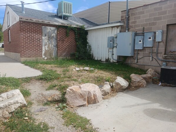 Un edificio de ladrillo con una unidad de aire acondicionado metálica en el techo, un camino de hormigón y una zona ajardinada con rocas.