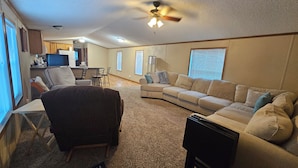 A living room with a sectional sofa, a dark armchair, a small table, and a ceiling fan. - Wonderful 3-bedroom mobile home with AC in Hyannis (Hyannis)