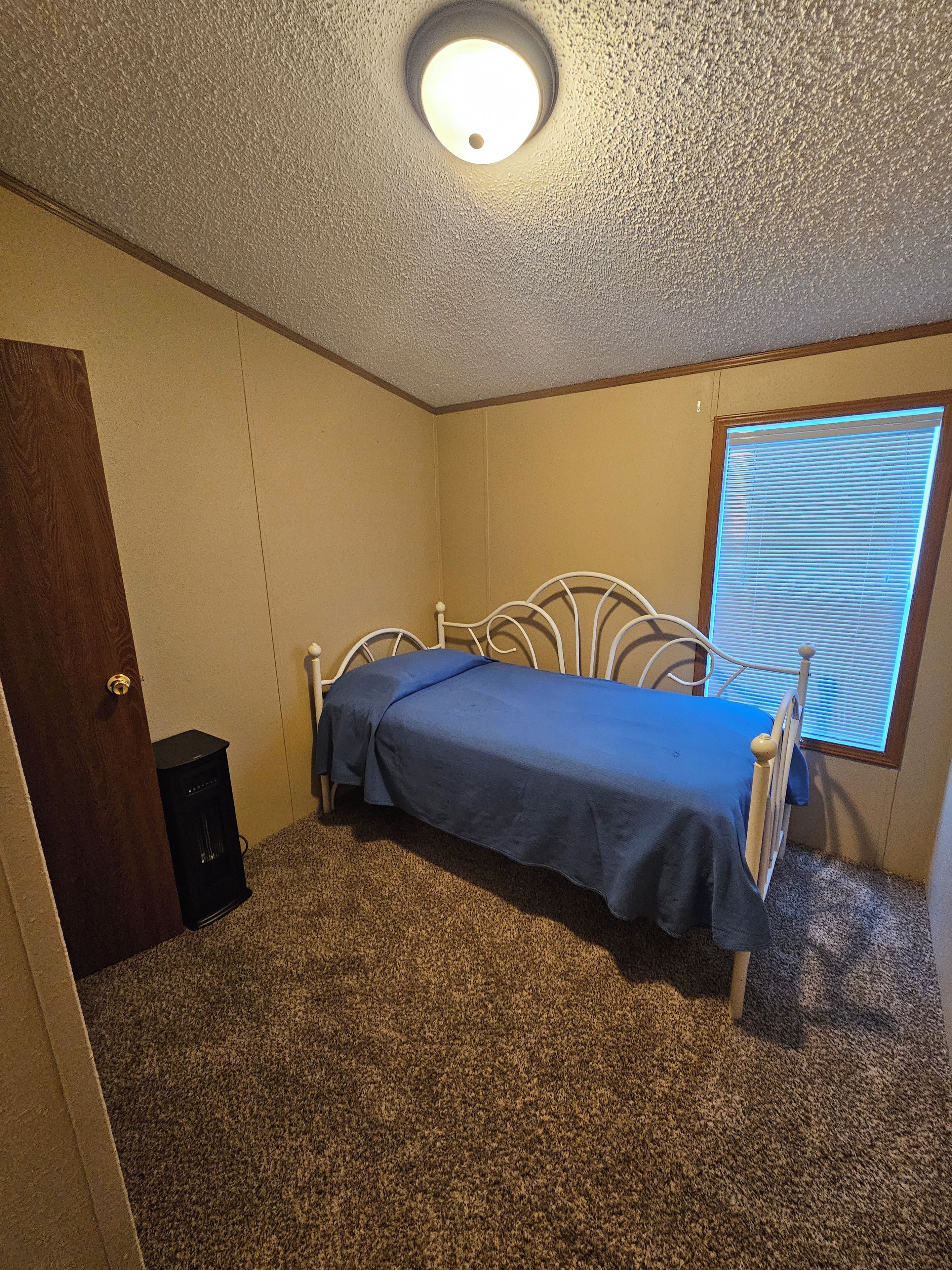 Ein Schlafzimmer mit einem Einzelbett, einer Tür, einem Fenster mit Jalousien und einem Teppichboden.