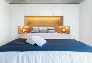 Quarto de hotel com uma cama grande, duas luminárias laterais e cabeceira de madeira.