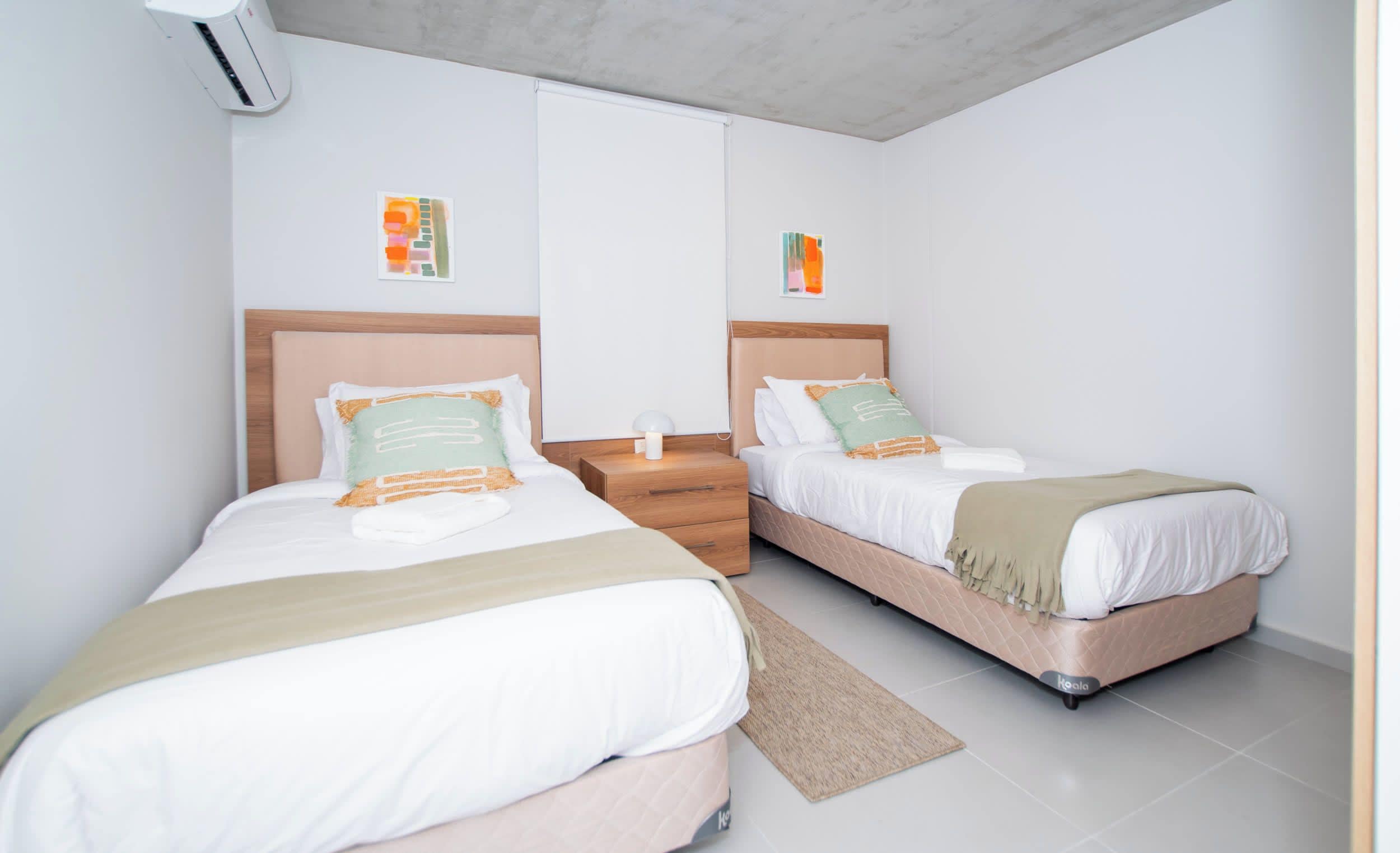 Habitación de hotel con dos camas, una mesita de noche, una lámpara y aire acondicionado instalado en la pared.