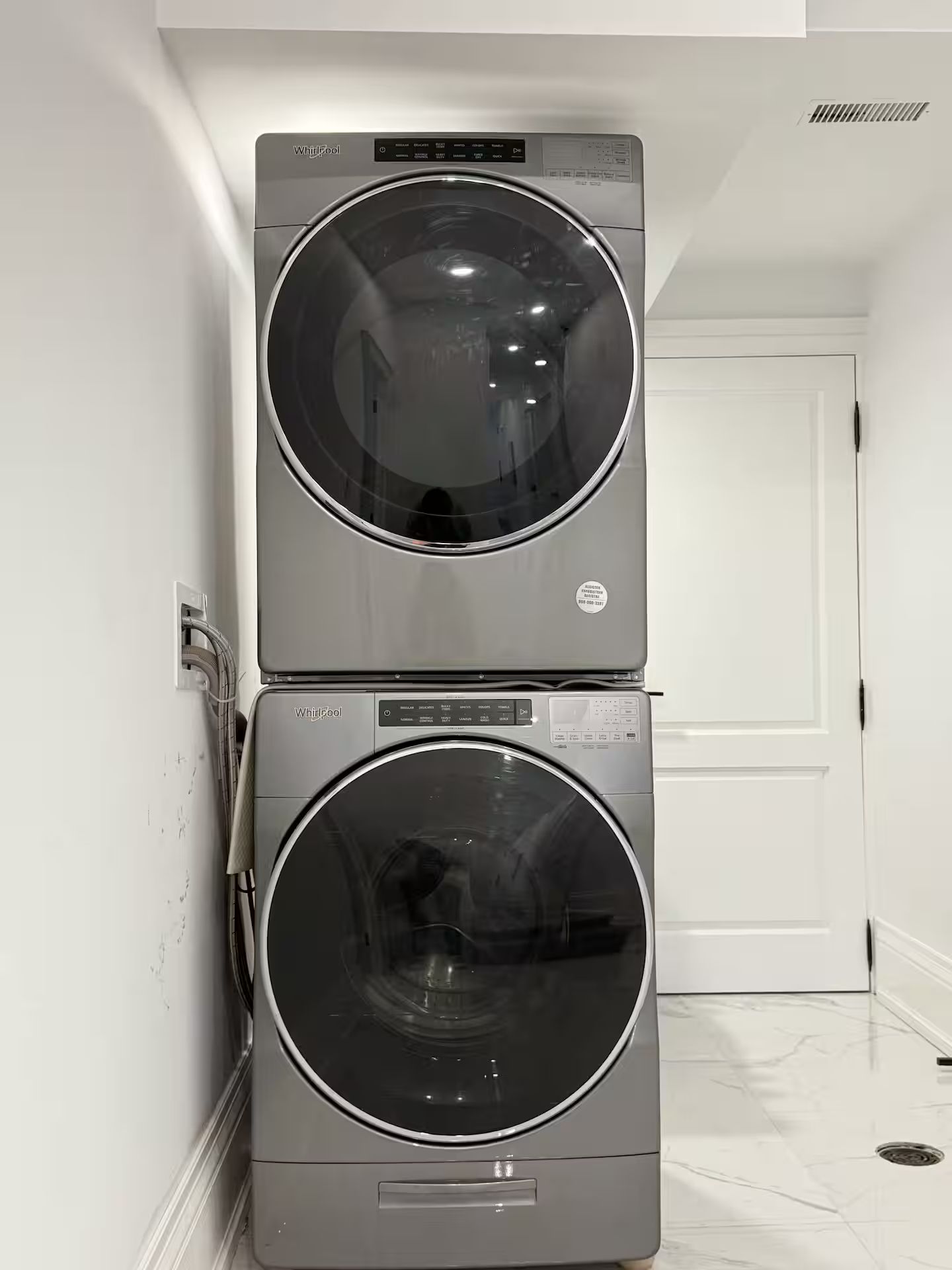Een gestapelde wasmachine en droger in een gang met een witte deur en een ventilatieopening in de muur.
