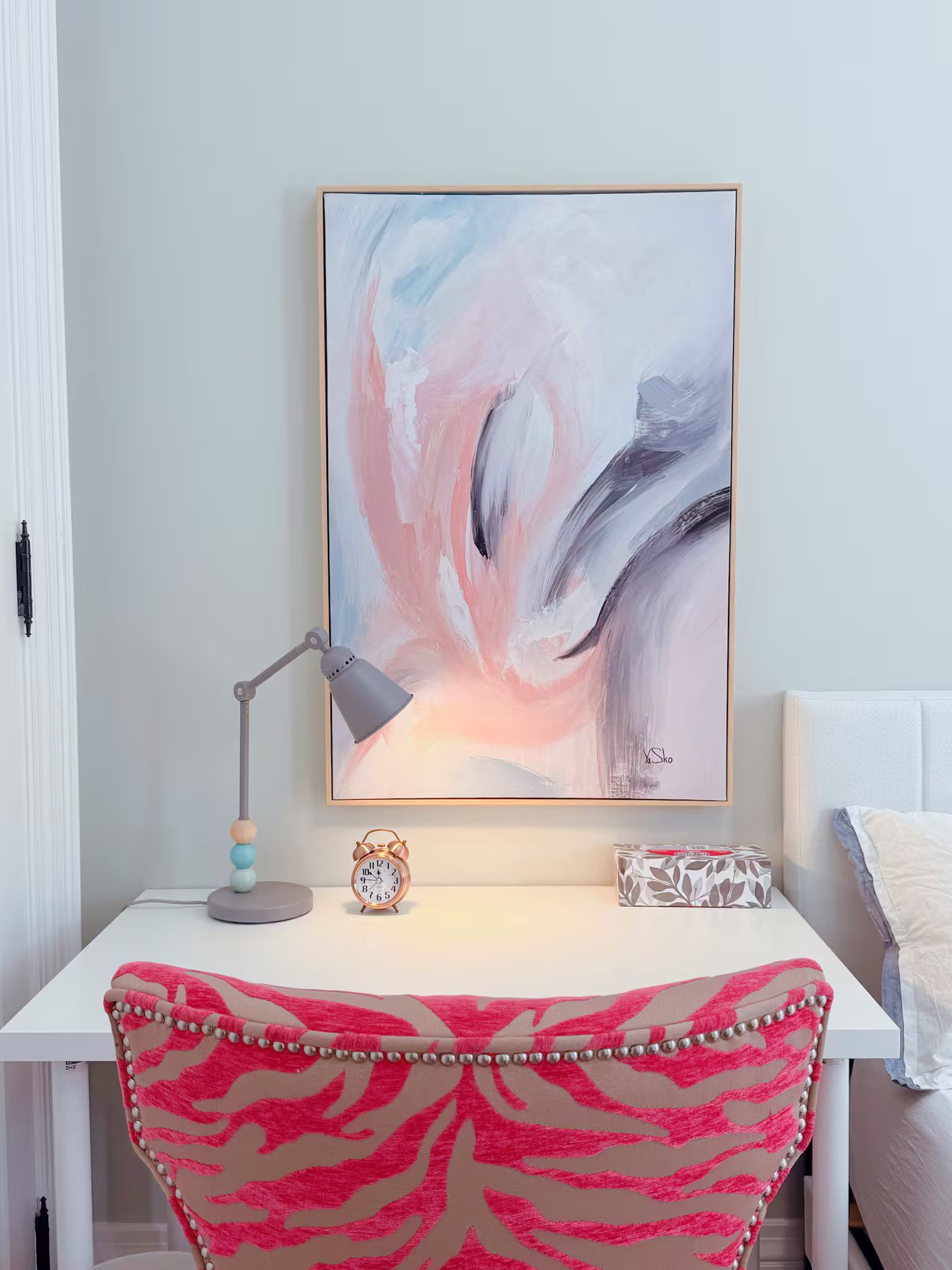 A room with a desk, a framed abstract painting, a desk lamp, a clock, and a chair with a pink and beige pattern.