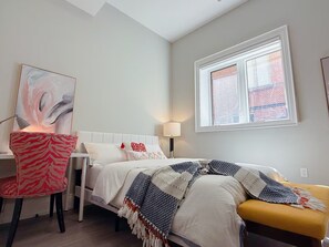 Een slaapkamer met een bed, een stoel, een lamp, een raam en een schilderij.
