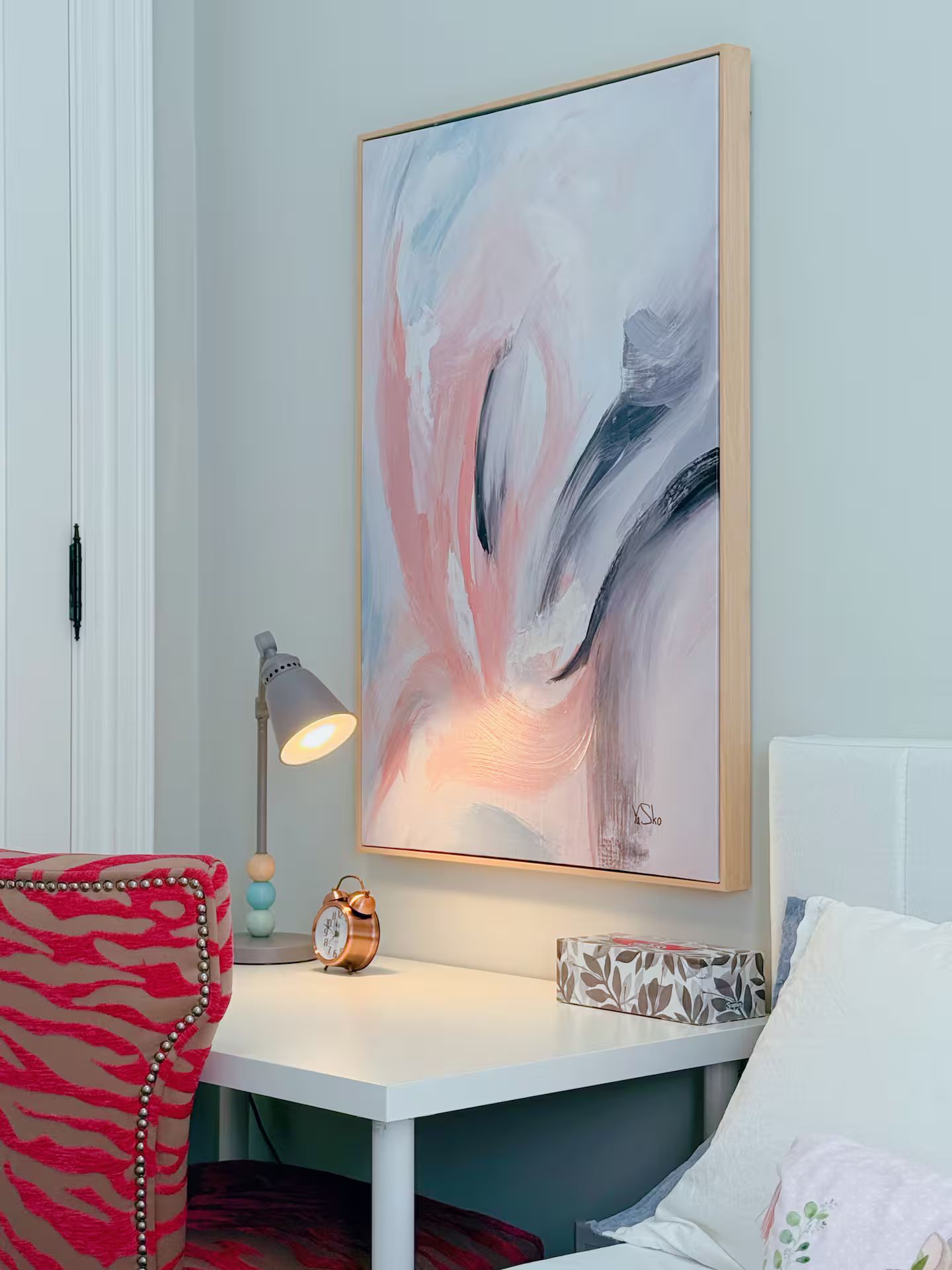 Een kamer met een wit bureau, een abstract schilderij in een lijst, een lamp, een klok en een kussen met dessin.