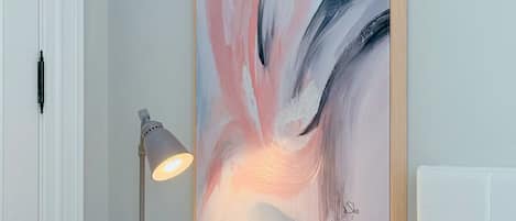 Una habitación con un escritorio blanco, un cuadro abstracto enmarcado, una lámpara, un reloj y un cojín estampado.