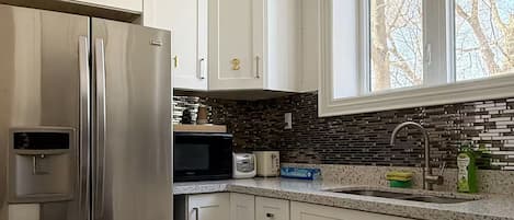 Una cocina con electrodomésticos de acero inoxidable, armarios blancos, encimera de granito y ventana con vista a árboles desnudos.