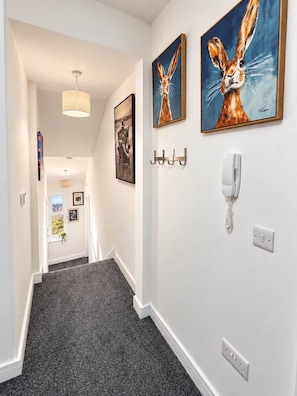 A hallway with a grey carpet, a white wall, and two framed paintings of rabbits. There are hooks on the wall and a white telephone. - King Bed • Free Parking • Stylish 1BR (Liverpool)