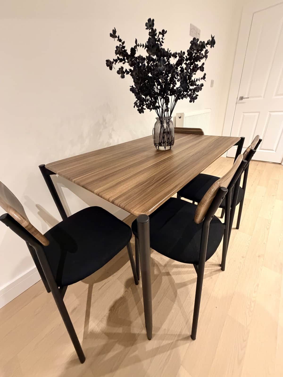 A dining table with a vase of flowers, four chairs, and a white door in the background.