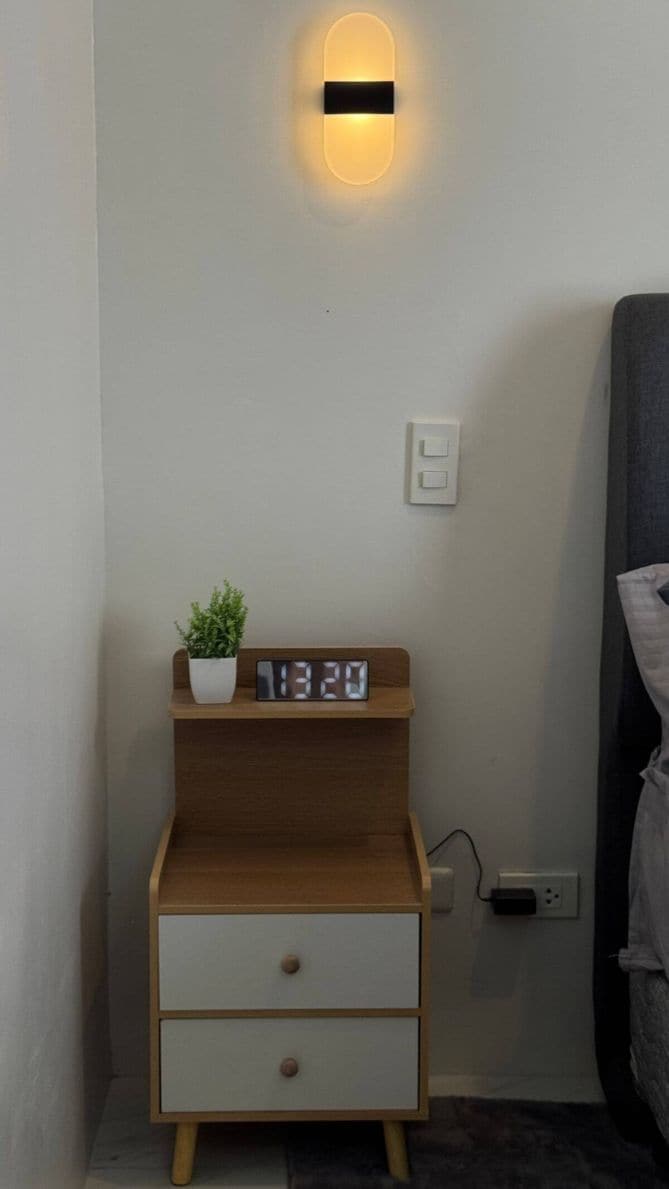 Een nachtkastje met een digitale klok, een klein plantje en een wandlamp.