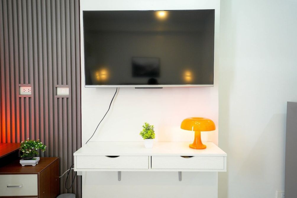 A modern room with a flat-screen TV mounted on the wall, a white console table with a small plant and an orange lamp, and a wooden cabinet with a plant on top.