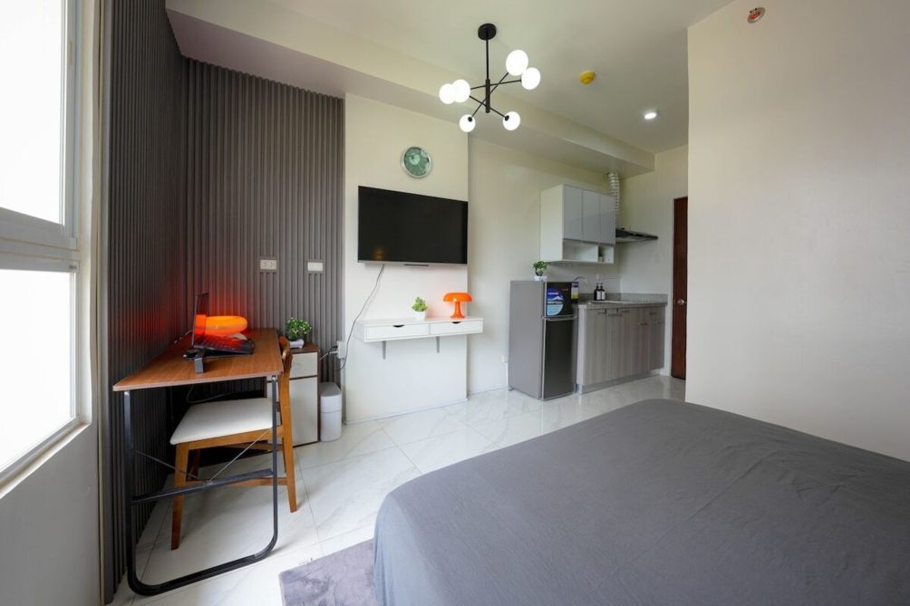 Ein modernes Schlafzimmer mit einem großen Bett, einem Schreibtisch mit Stuhl, einem Fernseher und einer Küchenzeile im Hintergrund.