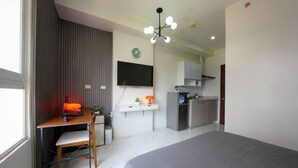 Quarto moderno com uma cama grande, uma mesa de escritório com cadeira, televisão e uma kitchenette ao fundo.