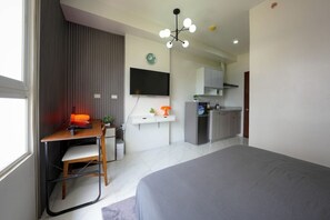 Ein modernes Schlafzimmer mit einem großen Bett, einem Schreibtisch mit Stuhl, einem Fernseher und einer Küchenzeile im Hintergrund.
