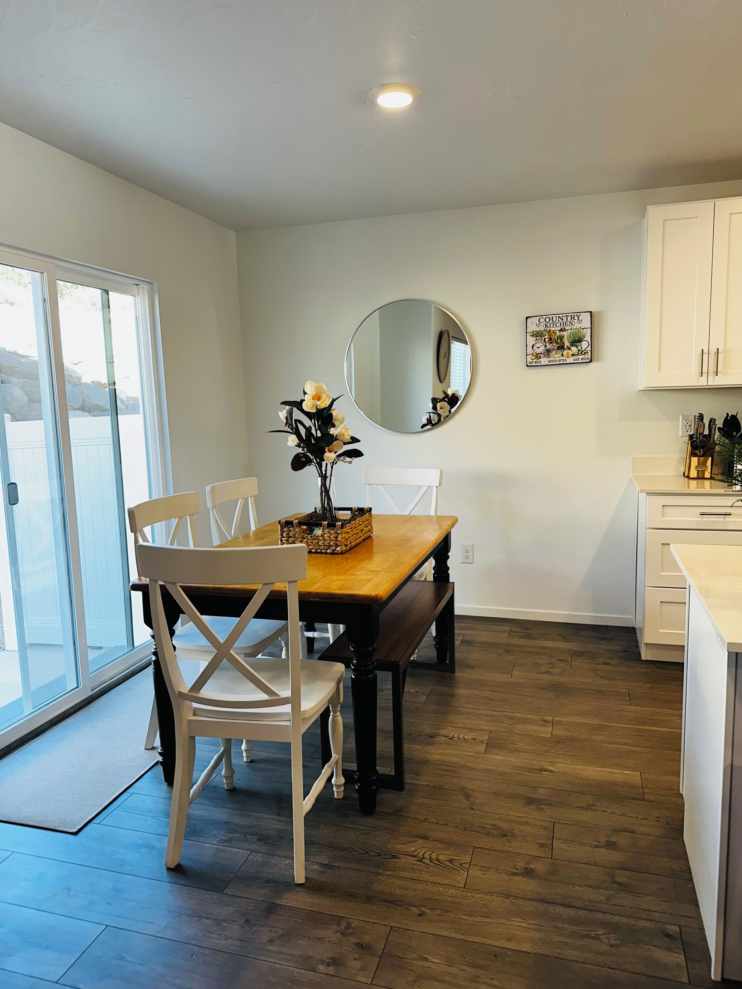 A dining area with a wooden table and chairs, a mirror on the wall, and a kitchen area with cabinets.