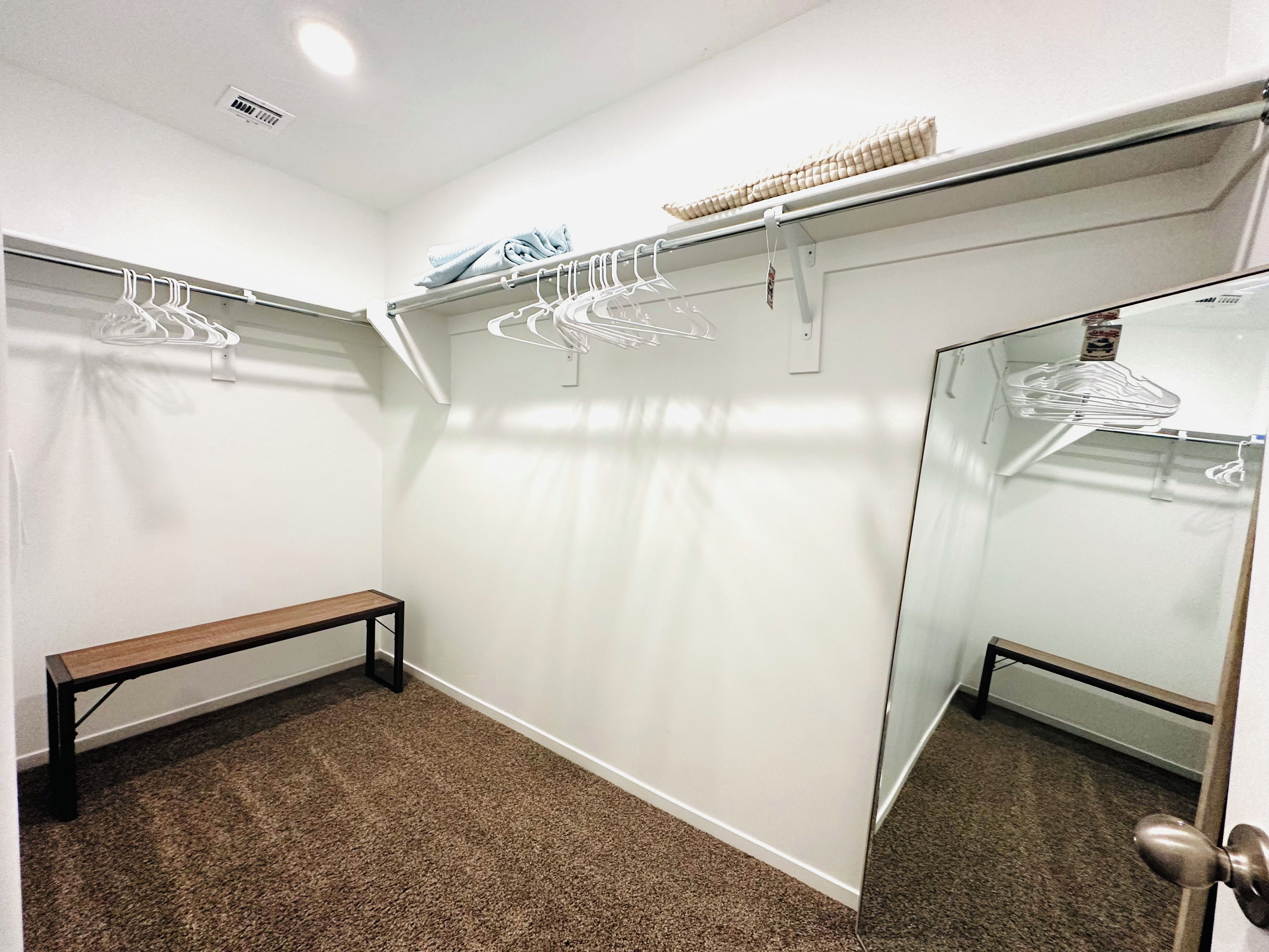 A walk-in closet with a bench, hanging clothes, and a mirror.