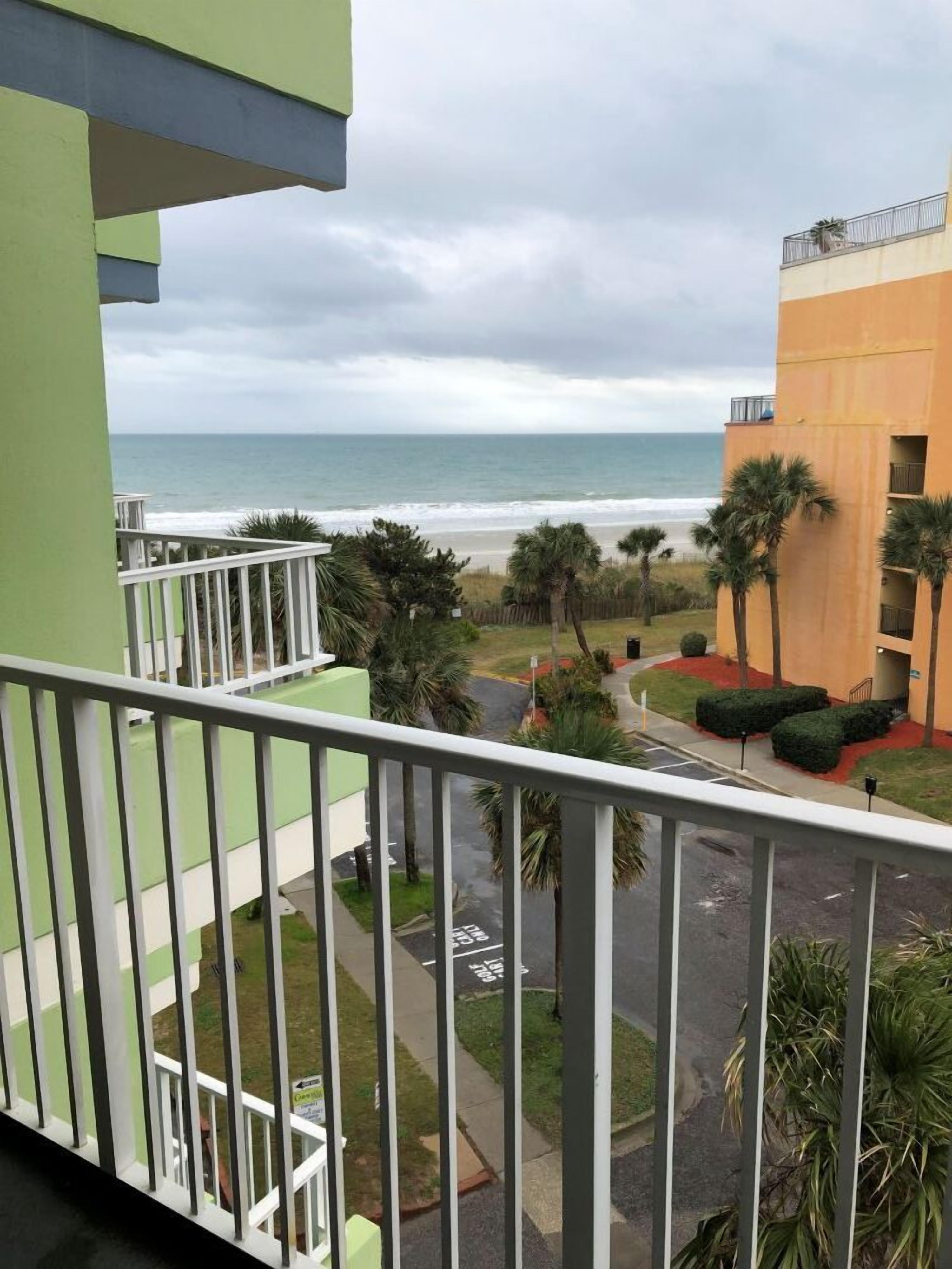 A balcony with a view of the ocean and a building with a palm tree garden.