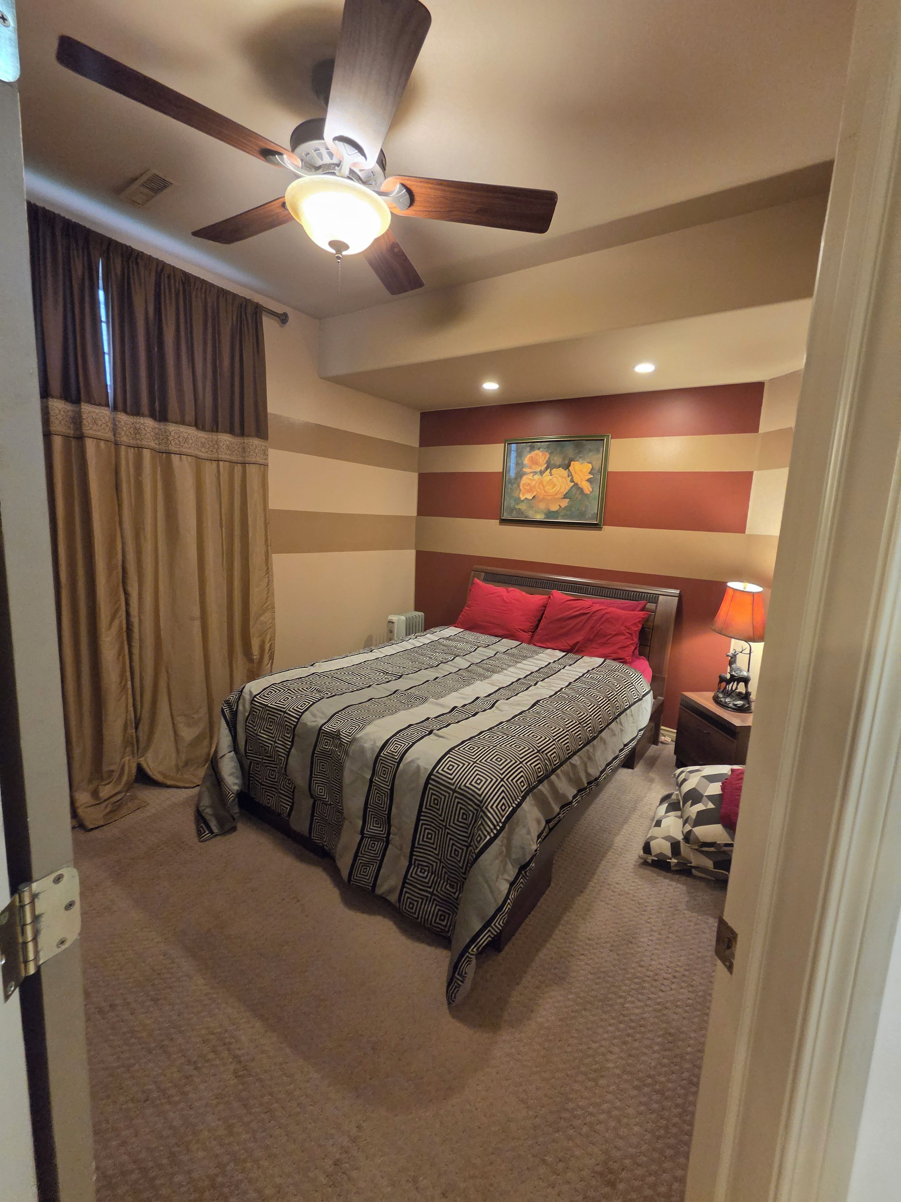 A bedroom with a bed, a ceiling fan, a radiator, a painting, and a nightstand with a lamp.