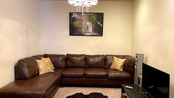 A living room with a brown leather sectional sofa, a mounted flat-screen TV, a chandelier, and a framed landscape painting on the wall.