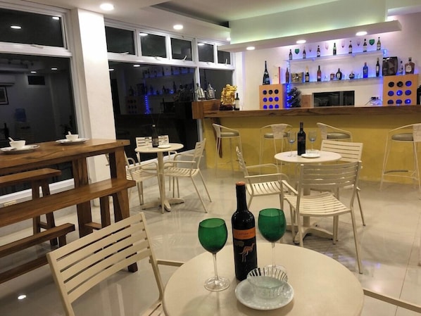 Un restaurante con mesas y sillas redondas, una zona de bar con estantes llenos de botellas, y una botella de vino sobre una mesa.