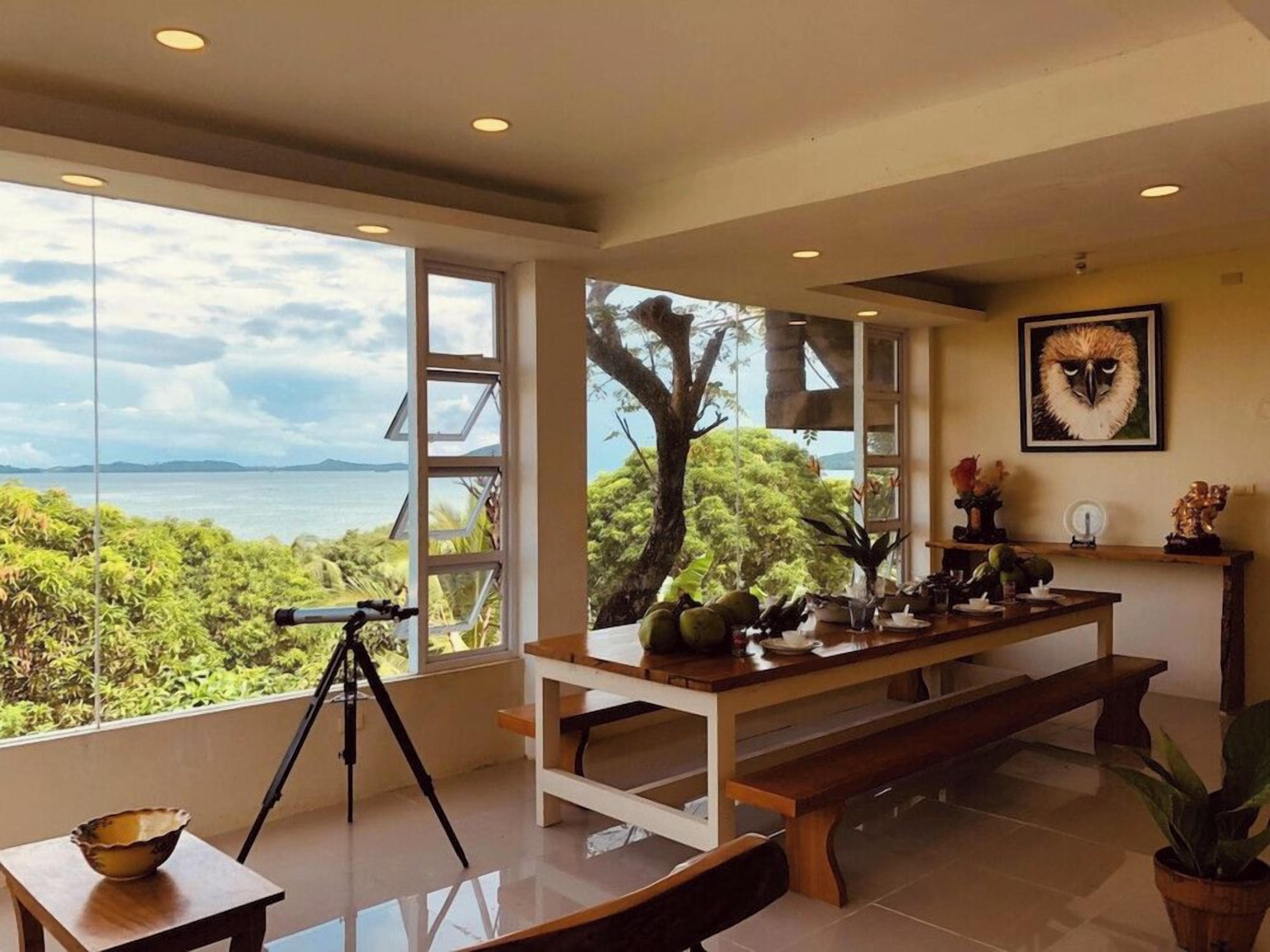 Une pièce avec un télescope monté sur un trépied, une table à manger avec un banc, et une vue sur la mer grâce à de grandes fenêtres.