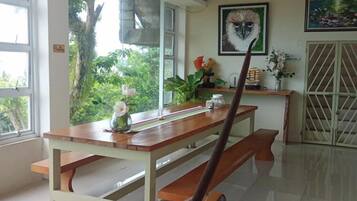 Un coin repas avec une table et des bancs en bois, une fenêtre donnant sur un paysage verdoyant et une porte surmontée d’un portail en métal.