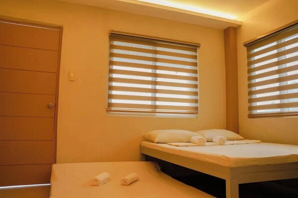 Una habitación con dos camas, cada una con una almohada y toallas enrolladas. Hay dos ventanas con persianas y una puerta.