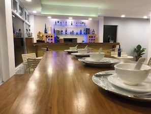 Un comedor con una larga mesa de madera dispuesta para cuatro, una barra con varias botellas y una planta a la derecha.