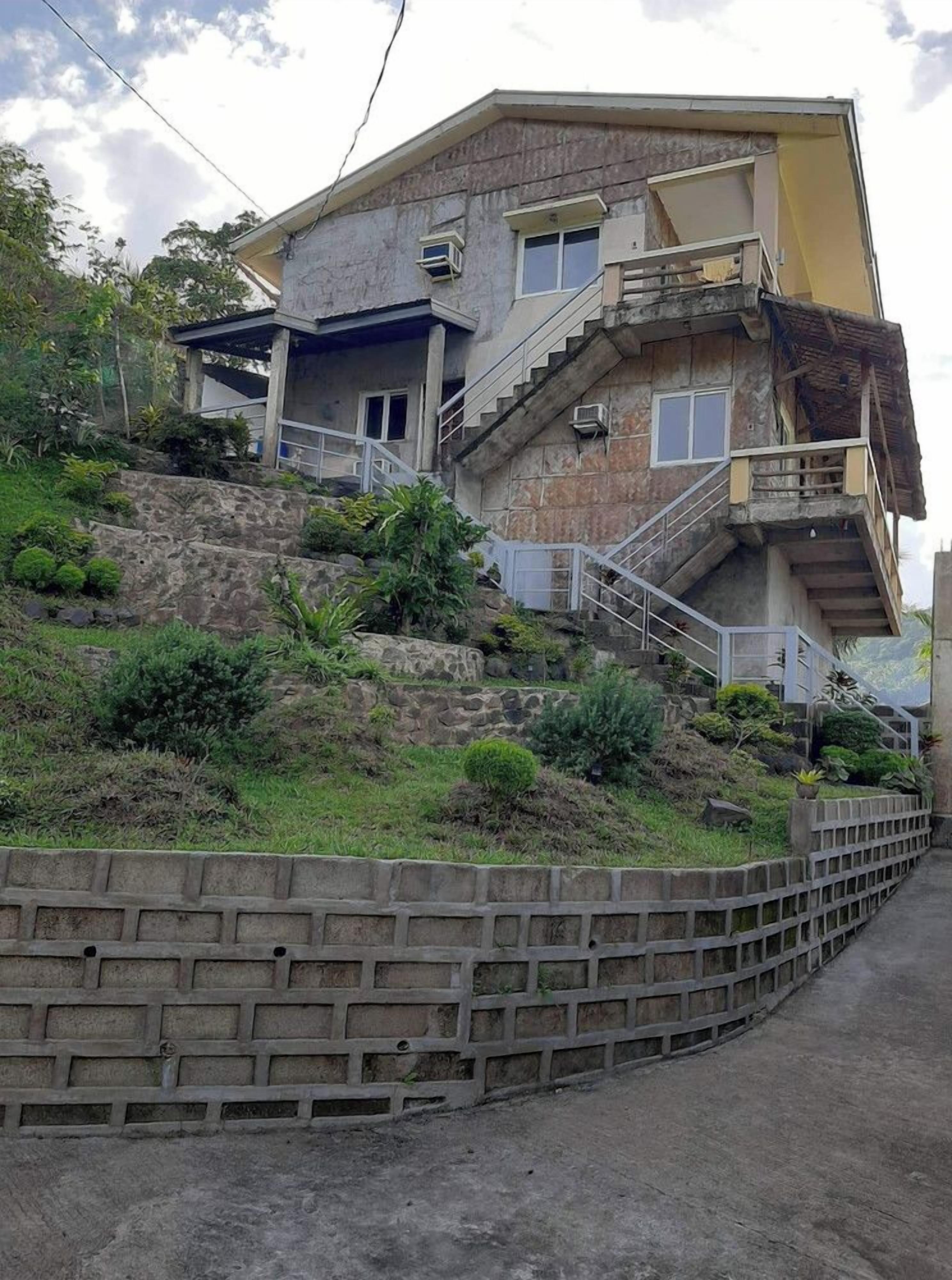 Un immeuble à plusieurs étages, doté d’un escalier menant à l’étage supérieur, entouré de verdure et bordé d’un mur de soutènement.