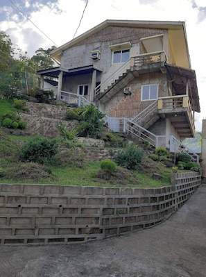 Un edificio de varios pisos con una escalera que conduce al nivel superior, rodeado de vegetación y un muro de contención.