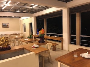 Un área de comedor con mesas y sillas de madera, un centro decorativo y ventanas amplias.