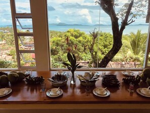 Une table à manger dressée avec des assiettes, des bols et un vase, donnant sur un paysage tropical avec vue sur la mer et les montagnes.