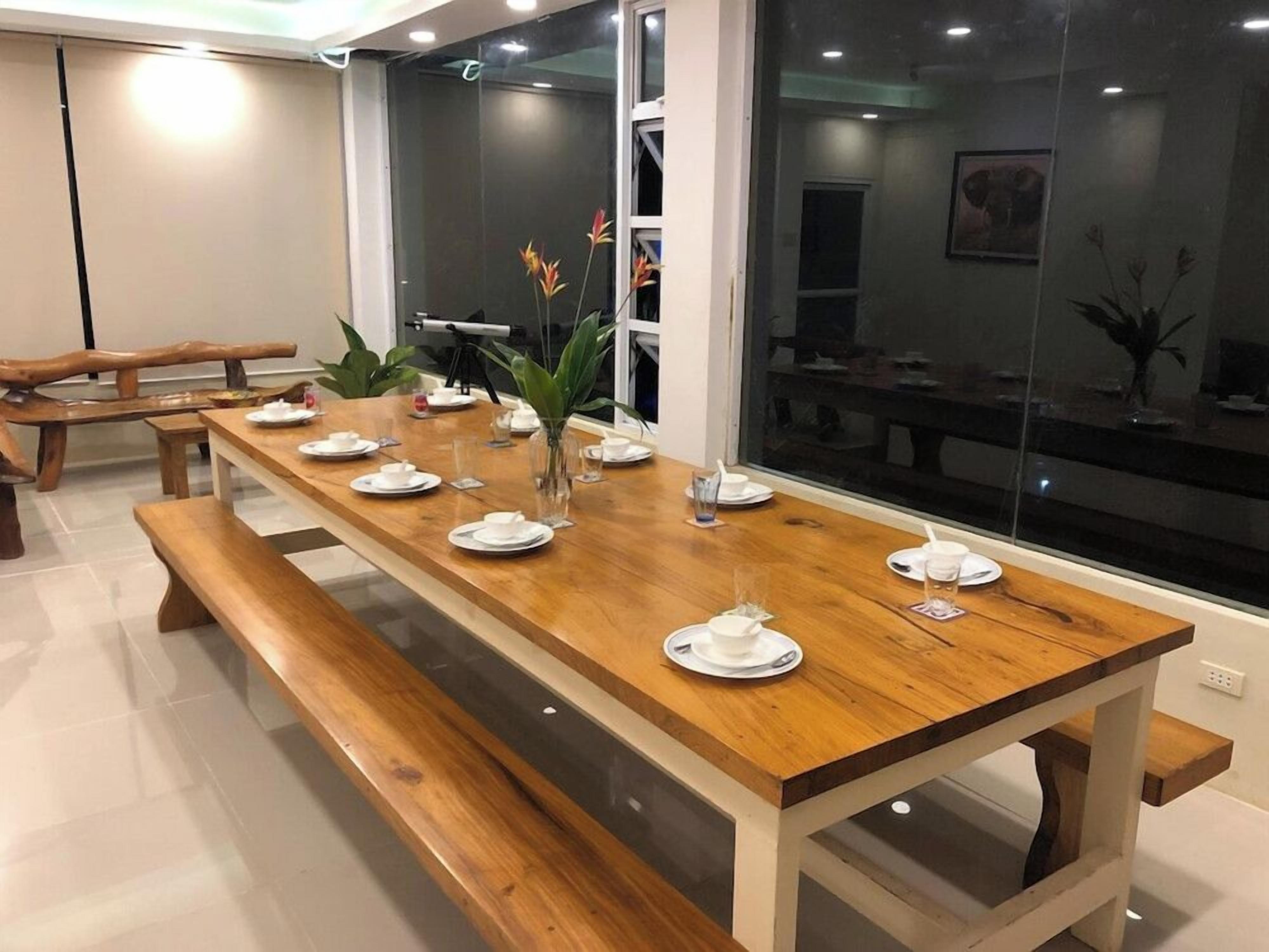 Un espace repas avec une longue table en bois dressée pour le repas, des bancs et une vue dégagée grâce à de grandes fenêtres.