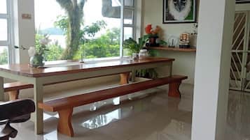 Een ruime kamer met een grote houten tafel en banken, een raam met uitzicht op groen, en een kunstwerk in een lijst aan de muur.
