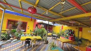 Un área de comedor al aire libre con mesas y sillas amarillas, rodeada de plantas en macetas y un piso de ladrillo.