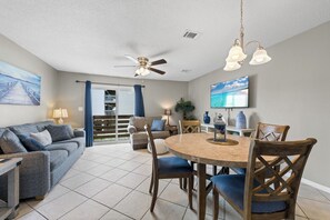 A living room with a sofa, a dining table with chairs, a ceiling fan, and a TV mounted on the wall. - Gulf View 8 with Pool (Gulf Shores)