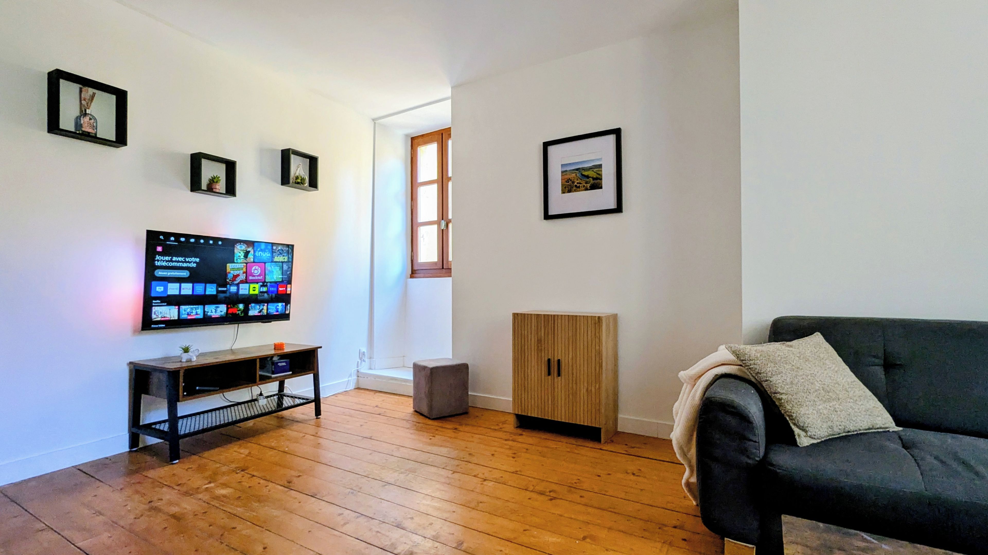 Un salon doté d’un téléviseur à écran plat, d’un parquet, d’un canapé foncé, d’une commode et de tableaux encadrés accrochés aux murs.