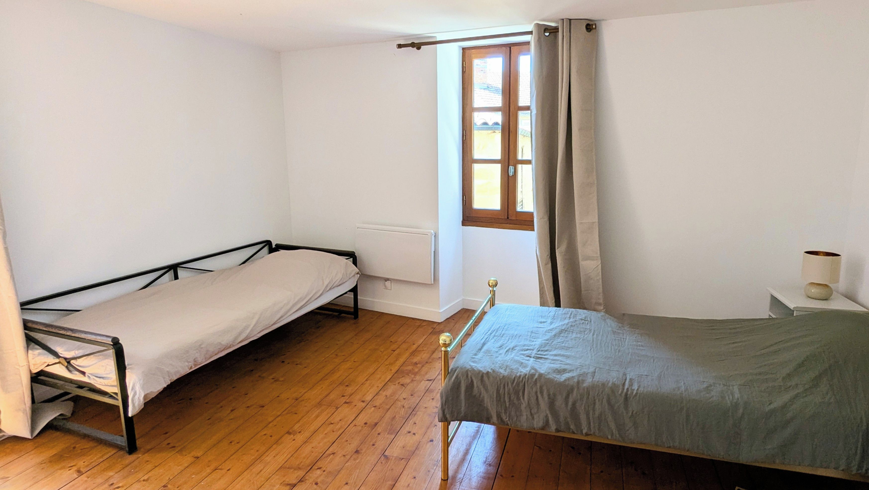 A room with two single beds, wooden flooring, a window with curtains, and a radiator.