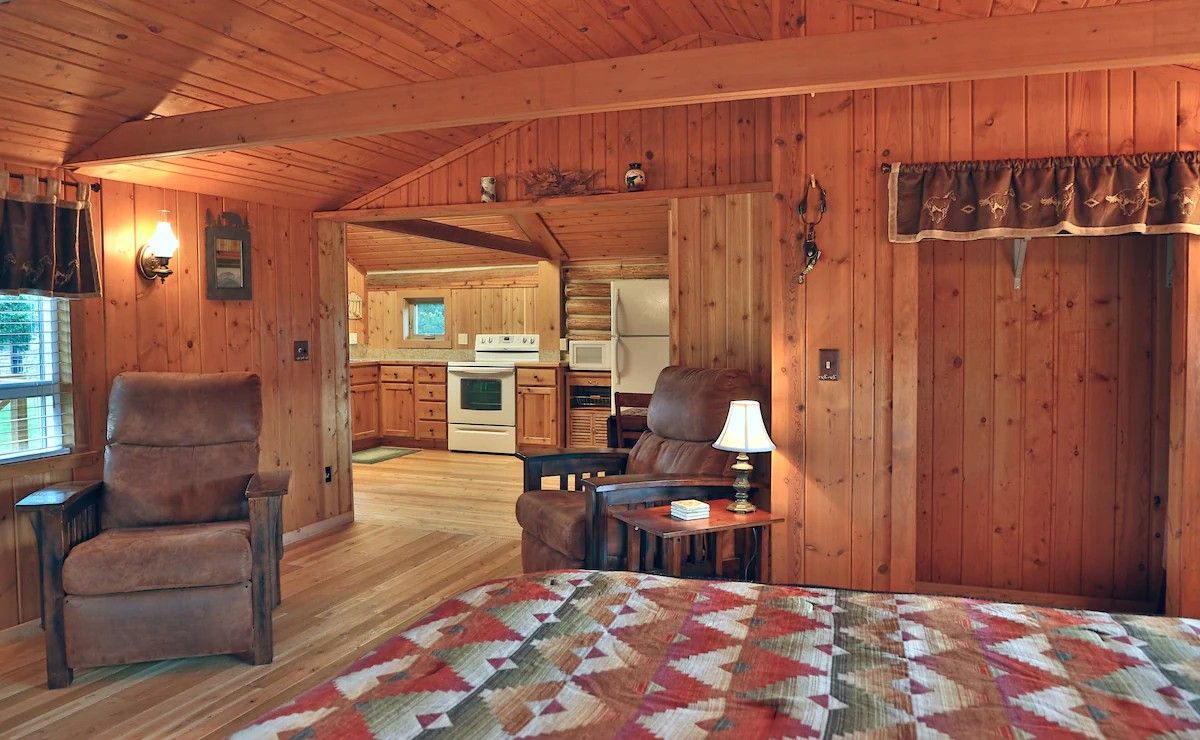A log cabin interior with a cozy living area, a dining table, and a kitchen visible through an open doorway.