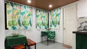 Um pequeno espaço fechado com teto de madeira, uma mesa verde com um banco e uma pequena mesa com uma planta.