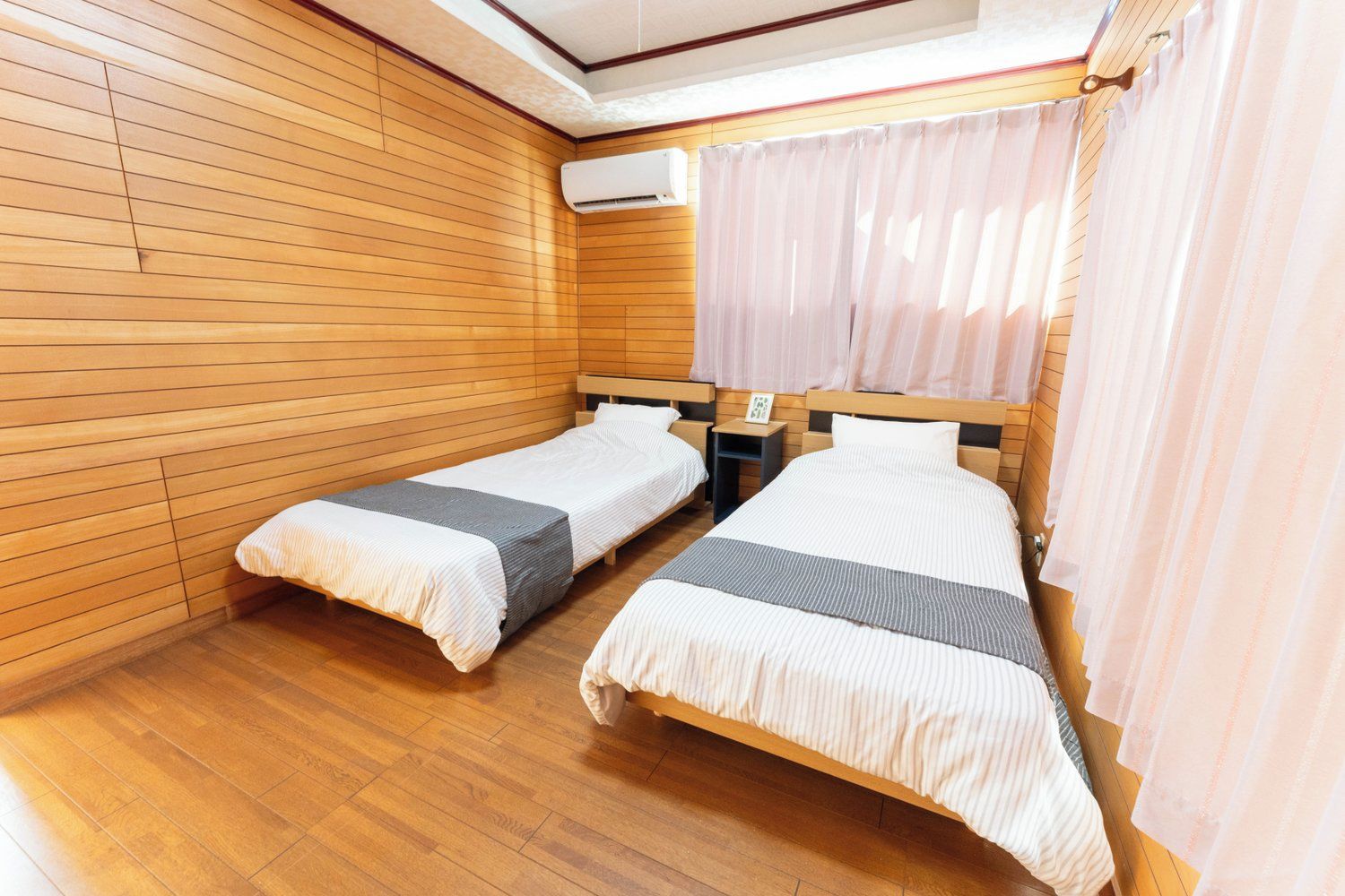 Une chambre avec deux lits simples, des murs lambrissés, un ventilateur de plafond et une fenêtre avec des rideaux.