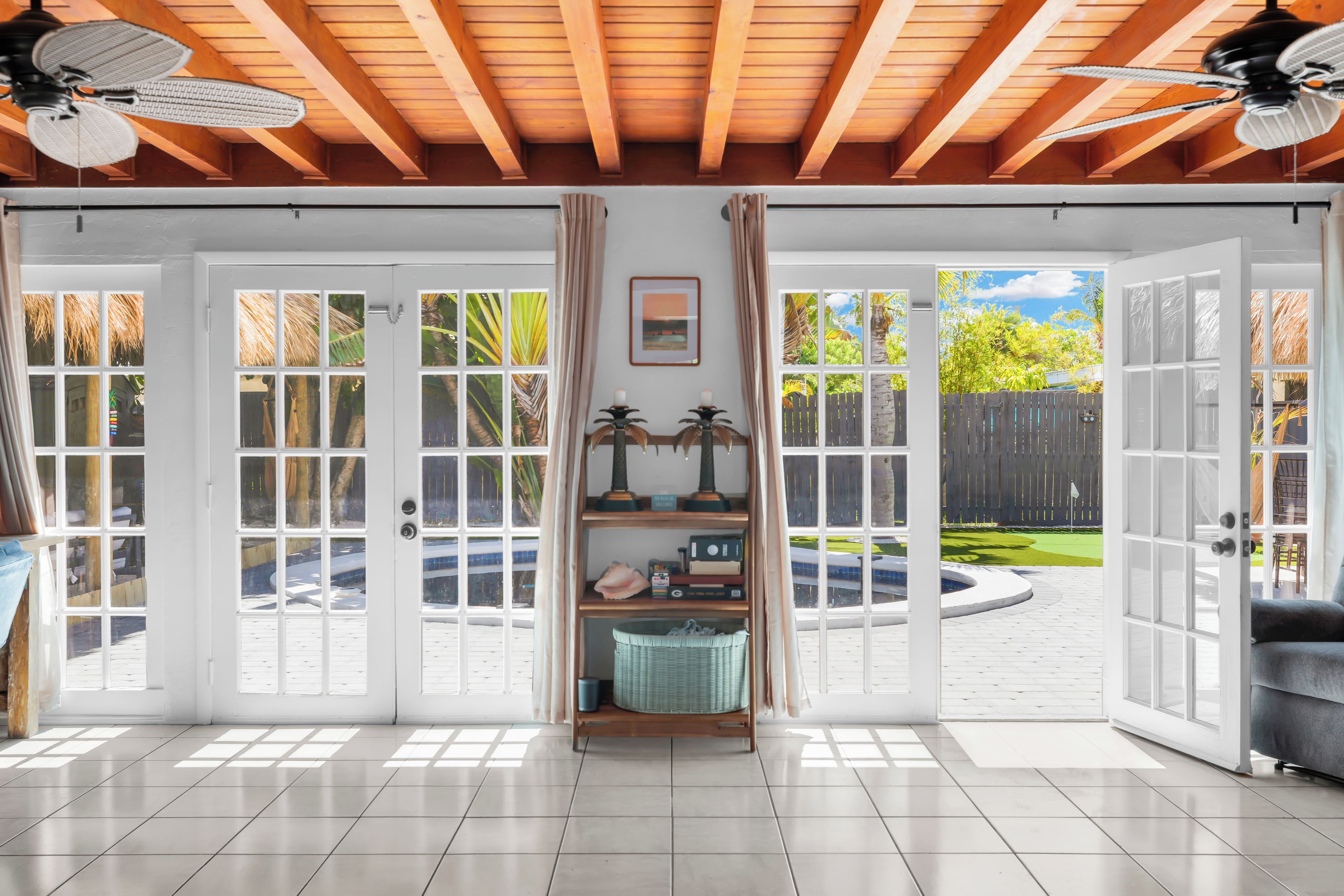 A spacious room with a tiled floor, a ceiling fan, and a view of a backyard with a pool and greenery.