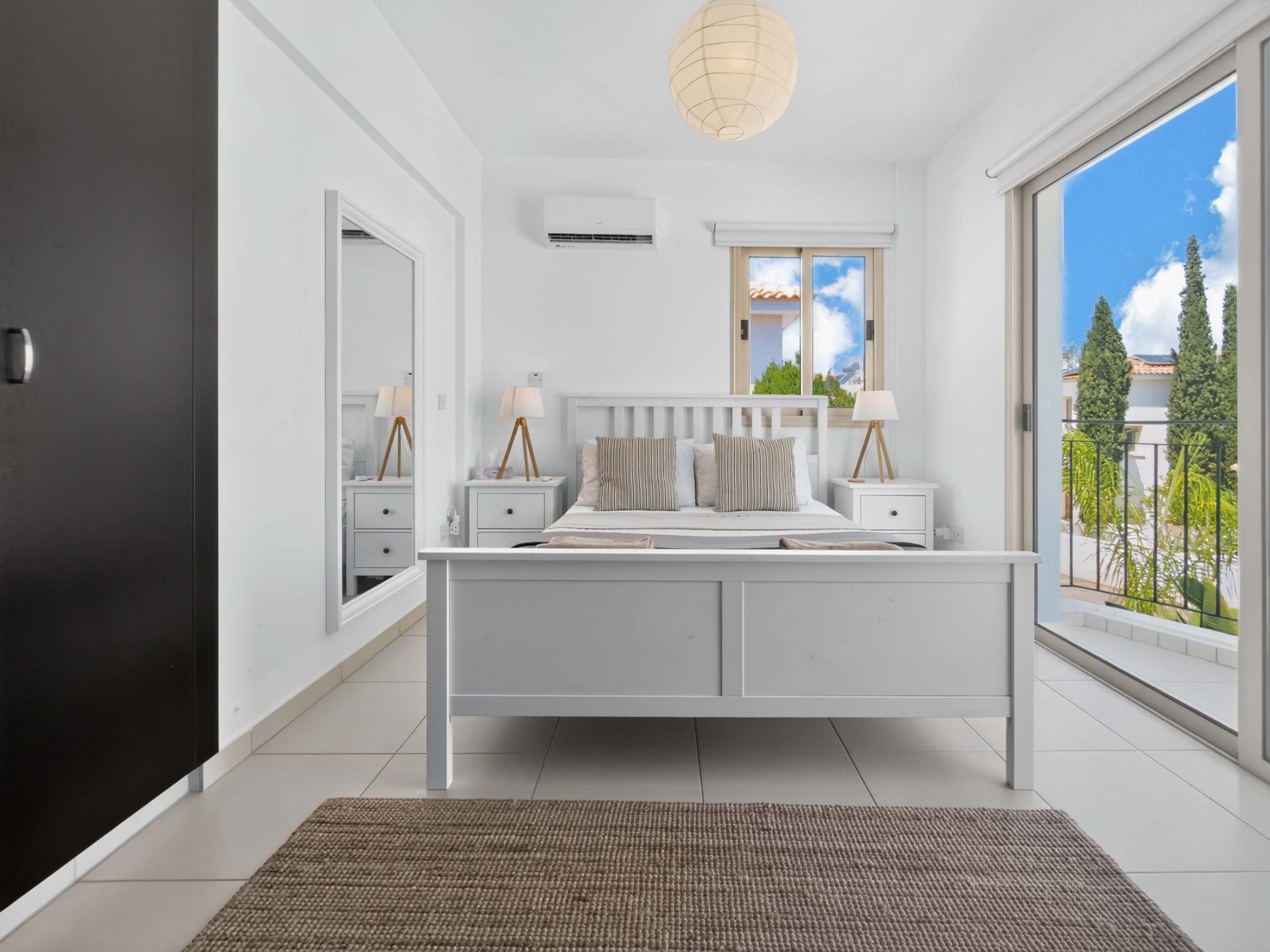 A modern bedroom with a white bed, bedside tables, a large window with a view, and a ceiling light fixture.